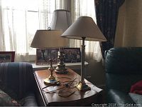 Photo of two table lamps on wooden side table, one brass with damaged shade, one metal with cream shade, cords and switches visible.