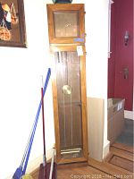 Full view of the grandfather clock standing against a wall showing the wooden case, glass panels, and pendulum chains inside.
