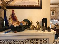 Resin rooster, brass tone ducks, tin houses, and metal candle holder on white shelf from left side view.