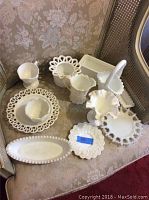 Photo of assorted milk glass items on a chair, featuring several vases, bowls, and footed cake plate
