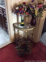 Demi lune glass top table with three large artificial flower arrangements on and one small at base, showing placement and scale.