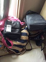 Photo showing a pile of assorted purses including a pink leather purse, black leather purse, navy blue and cream woven tote, and dark gray backpack.