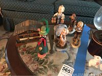Image shows a variety of Hummel figurines including a small wooden house, a figure wearing a green hat with a feather, and several child figurines arranged on a glass table.