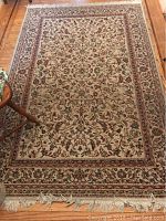 Full view of rectangular beige area rug with maroon and green floral and vine design, fringed edges laid on a wooden floor.
