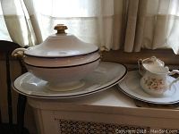 Wide angle photo of the tureen on its serving platter next to sugar bowl and milk pitcher, shows porcelain items with wear and gold trim