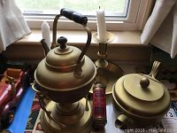 Antique brass teapot with black handle and lid, vintage brass teapot on warmer stand, and brass candlestick with candle on table by window