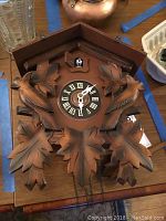 Full front view of vintage German cuckoo clock showing wooden carved leaves and birds, clock face with Roman numerals, cuckoo bird above clock face, chains hanging below without weights.