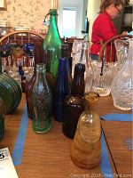 Photo showing a collection of vintage glass bottles on a wooden table with various colors and shapes, including green, brown, blue, and amber bottles and some clear glass decanters.