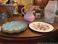 Image showing all items on a wooden table: two blue milk glass egg platters with gold trim, Johnson Brothers floral plate, pink pitcher, and other items in background.