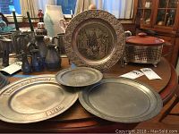 Four pewter plates on a wooden table, one with intricate winter village scene and one marked Wilton Columbia PA