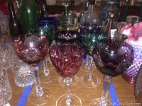 Set of nine vintage colored long stem cut glass wine glasses on a table seen from above, showing variety of colors including purple, green, blue, pink.