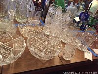 Overview of various leaded cut glass bowls, vases, and glasses arranged on a wooden table showing intricate cut patterns.