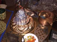 Various copper molds, a copper pot, and a copper tea kettle displayed on a table along with silver plate trivets and decorative items.