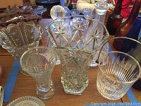 Multiple clear cut glass items on wooden table including a large ice bucket and different vases with various cut patterns