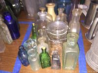 Various antique and vintage glass bottles in assorted colors and shapes, including clear, green, and blue glass, some with embossed text and a glass jar with lid.
