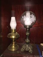 Two electric hurricane lamps on a wooden surface, one brass based with frosted chimney globe and the other ornate metal based with floral decorated clear globe.