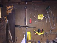 Wall-mounted pegboard with multiple vintage hand saws hung by their wood handles, a small hand saw, and a hand drill. Also visible are yellow tool packs and small hand tools.