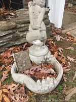 Full view of cement fountain sculpture of little girl holding watering can on base bowl, showing cracks on bowl edge and leaves on the ground around it.