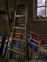 Photo of aluminum A-frame ladder standing near RE/MAX real estate signs inside a basement, shows overall height and side view.