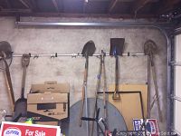 Photo of yard tools hung on a wall-mounted rack including shovels and a hoe, with cardboard boxes and a folding table stored below.