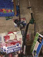 Photo showing FeatherLite gas weed eater with real estate signs in background, tools arranged upright against wall.