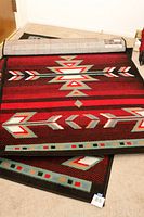 Two Native American style area rugs partially rolled and laid on carpet floor, showing front patterned surfaces and part of backing.