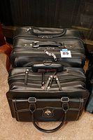 Two black leather laptop roller bags stacked, showing multiple zippered compartments and handles.
