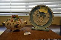 Wide photo showing two items on wood surface in front of window blinds: the blue Inoclay Studios collectors plate with white relief figure scene on stand and the ornate multicolored floral Capodimonte footed bowl with lid.