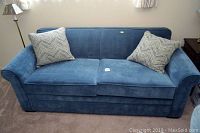 Front view of emerald blue sleeper sofa with two decorative pillows on beige carpeted floor, lit by natural window light