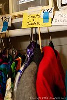 View of multiple coats and jackets hanging on hangers on a rod with sign 'Coats & Jackets' and lot number 132 visible.
