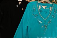 Turquoise top with bead and sequin floral design next to black jacket with silver buttons on hanger