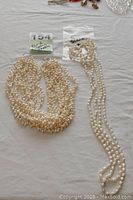 Two freshwater pearl necklaces on a white surface, one being an 8 strand choker and the other a single strand opera length necklace in sealed plastic bag