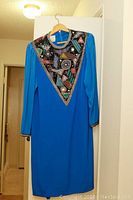 Full length photo of the bright blue dress hanging on wooden hanger showing large beaded triangular collar and long sleeves
