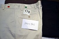 Close-up photo of beige men's casual pants size 34x30 with belt loops and buttoned back pocket, marked with red and green tags on waistband.