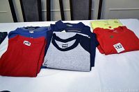 Nine folded men's casual shirts in various colors including red, blue, gray, yellow, and navy, shown on a table backdropped by chair backs.