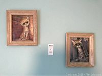 Two 'Pity Kitty' paintings by Gig hanging on a wall, framed in natural wood-tone frames. Left painting shows a sad cat sitting by a brick wall, right shows a sad cat in a wooden enclosure.