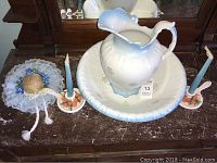 Wash set displayed on dark surface: white semi porcelain pitcher with partial crack and scalloped bowl with blue tint and floral motifs. Also seen are blue candles on floral holders and a lace doily with decorative ball.
