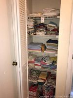 Photo of linen closet shelves showing folded bath towels, kitchen towels, table linens, aprons, and light bulb boxes organized on wire shelving.