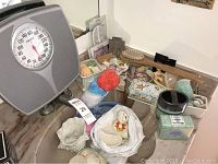 Photo of assortment of bathroom seashell items displayed on countertop including a gray mechanical bathroom scale, porcelain seashell figurine, small baskets, various soaps, loofah sponges, and decorative boxes.