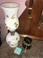 Full view of the antique white glass hurricane lamp next to the broken decorative dish with black and gold design.