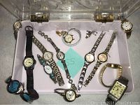 Top-down photo of eleven assorted analog watches laid out in a clear plastic container with purple padding beneath them