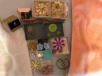 Photo showing assorted brooches mostly floral designs and two pairs of earrings arranged on a peach and gray fabric surface, some in boxes.