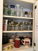 Photo showing kitchen cupboard with several shelves of glass jars, bottles, and assorted mugs and cups