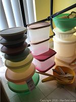 Photo showing stacked round and rectangular Tupperware containers in various colors with lids, plus yellow utensils on table.