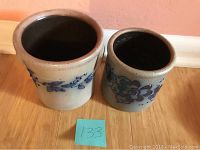 Two crocks side by side on hardwood floor, one larger with blue floral decoration, one smaller plain with blue flower motif.