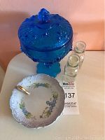 Blue glass candy dish with lid and plate shown on table, with two small clear glass bottles beside them