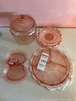 Collection of pink Depression glass items including lidded cookie jar, relish dish, and crocheted doilies on table.