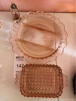Top view of two pink glass relish dishes placed on a table. One is round with scalloped edges and divided into three compartments with a starburst pattern. The other is rectangular with textured geometric patterns and handles.