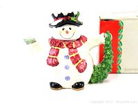 Photo of the snowman teapot showing front view highlighting the spout shaped as an arm and the tree handle, next to original box with festive design.