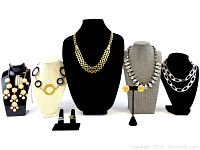 Front view of 5 vintage necklaces displayed on black and beige bust stands, 1 pair of yellow flower-shaped earrings on a stand, and 2 rings with green stones on black ring holders.